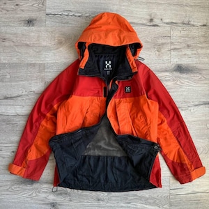 Puede incluir: Una chaqueta con capucha roja y naranja con forro de malla negra. La chaqueta tiene cierre de cremallera y dos bolsillos en la parte delantera. El nombre de la marca "Haglöfs" es visible en la parte delantera de la chaqueta.
