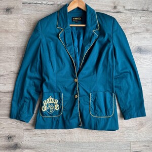 Puede incluir: Blazer color turquesa con ribetes y botones dorados. El blazer tiene dos bolsillos delanteros con detalles bordados en dorado. La marca "POMPOOS" es visible en la etiqueta. El blazer está colgado en una percha de madera.