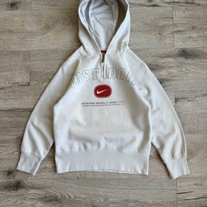 Op de afbeelding: Een lichtgrijs hooded sweatshirt met een rood Nike-logo en de tekst "Just Do It" in zwarte omlijnde letters. De tekst "Sporting Product Since 1971" is onder het logo gedrukt. Het sweatshirt heeft een ritssluiting.