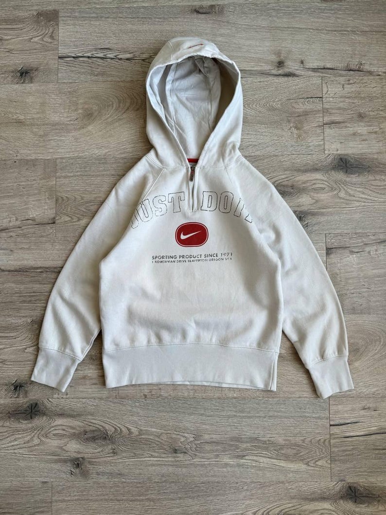 Op de afbeelding: Een lichtgrijs hooded sweatshirt met een rood Nike-logo en de tekst "Just Do It" in zwarte omlijnde letters. De tekst "Sporting Product Since 1971" is onder het logo gedrukt. Het sweatshirt heeft een ritssluiting.