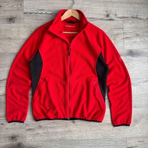 以下が含まれることがあります： 黒いサイドパネルとフルレングスのジッパーが付いた赤いフリースジャケット。 ジャケットはハイカラーと2つのフロントポケットを備えています。 ブランド名は胸に表示されています。 ジャケットは木製のハンガーに掛かっています。