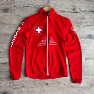 Puede incluir: Una chaqueta roja brillante con cremallera blanca y cuello alto. La chaqueta presenta un emblema de la cruz suiza en el pecho y la manga, con el texto "X BIONIC" en la manga izquierda. La chaqueta está colgada en una percha de madera.