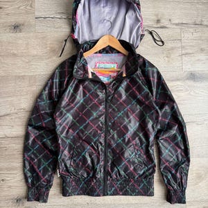 Puede incluir: Chaqueta negra con capucha y un estampado de cuadros coloridos. La chaqueta tiene cremallera completa, puños elásticos y capucha con cordón. El forro interior es gris claro. La etiqueta dice "Zoo York".