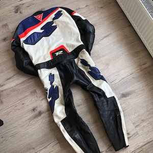 K&ouml;nnte beinhalten: Ein wei&szlig;er, blauer und roter Leder-Motorradrennanzug mit schwarzem Lederbesatz. Der Anzug hat einen Rei&szlig;verschluss am linken Bein und ein gro&szlig;es "R" auf dem R&uuml;cken.