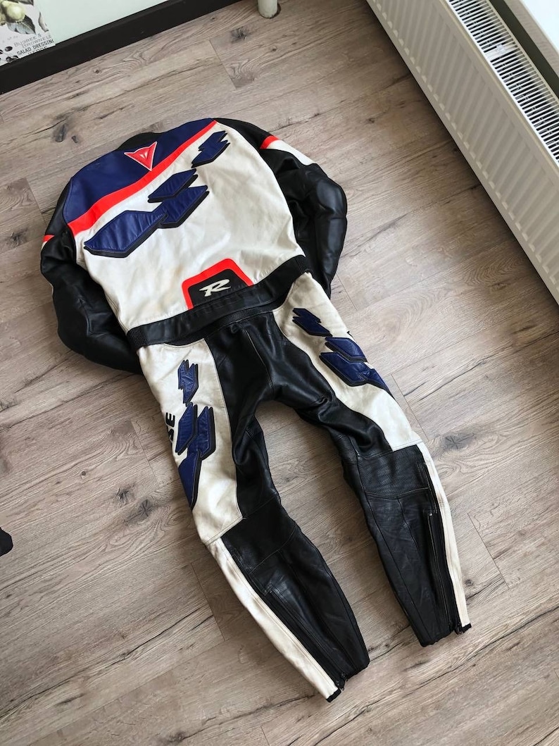 K&ouml;nnte beinhalten: Ein wei&szlig;er, blauer und roter Leder-Motorradrennanzug mit schwarzem Lederbesatz. Der Anzug hat einen Rei&szlig;verschluss am linken Bein und ein gro&szlig;es "R" auf dem R&uuml;cken.