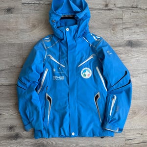 May include: A blue ski jacket with black and silver accents. The jacket has a hood and a zip closure. The jacket has a logo on the left chest that says "SHS" and a logo on the right chest that says "Monte Rosa".