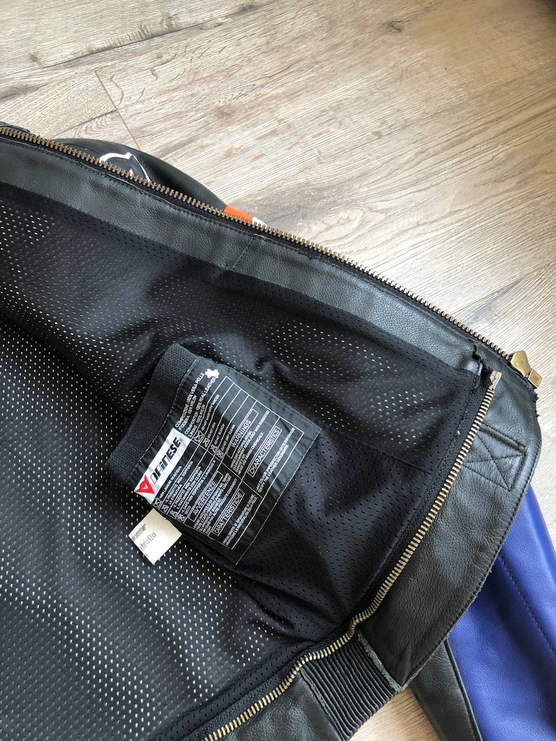 K&ouml;nnte beinhalten: Schwarze Leder-Motorradjacke mit einem blau-orangen Farbblock-Design. Die Jacke hat einen Rei&szlig;verschluss und ein Mesh-Futter. Das Etikett im Inneren der Jacke lautet "Dainese".