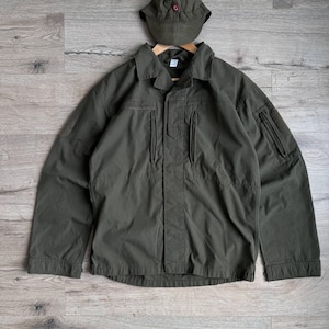 May include: Olive green jacket and matching cap. The jacket has a collared neckline, multiple zippered pockets, and a button-down front. The cap features a small red emblem. The set is displayed on a wooden surface.