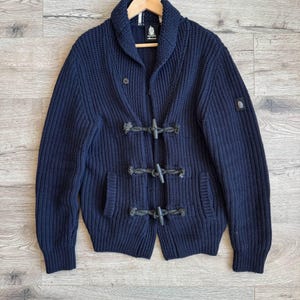 May include: A navy blue knit cardigan with a shawl collar and toggle closures. The sweater has two front pockets and a small logo patch on the left sleeve. The cardigan is hanging on a wooden hanger.