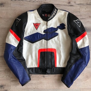 K&ouml;nnte beinhalten: Eine wei&szlig;e, schwarze und blaue Leder-Motorradjacke mit roten Akzenten. Die Jacke hat einen Rei&szlig;verschluss und ein Dainese-Logo auf der linken Schulter und dem rechten &Auml;rmel.