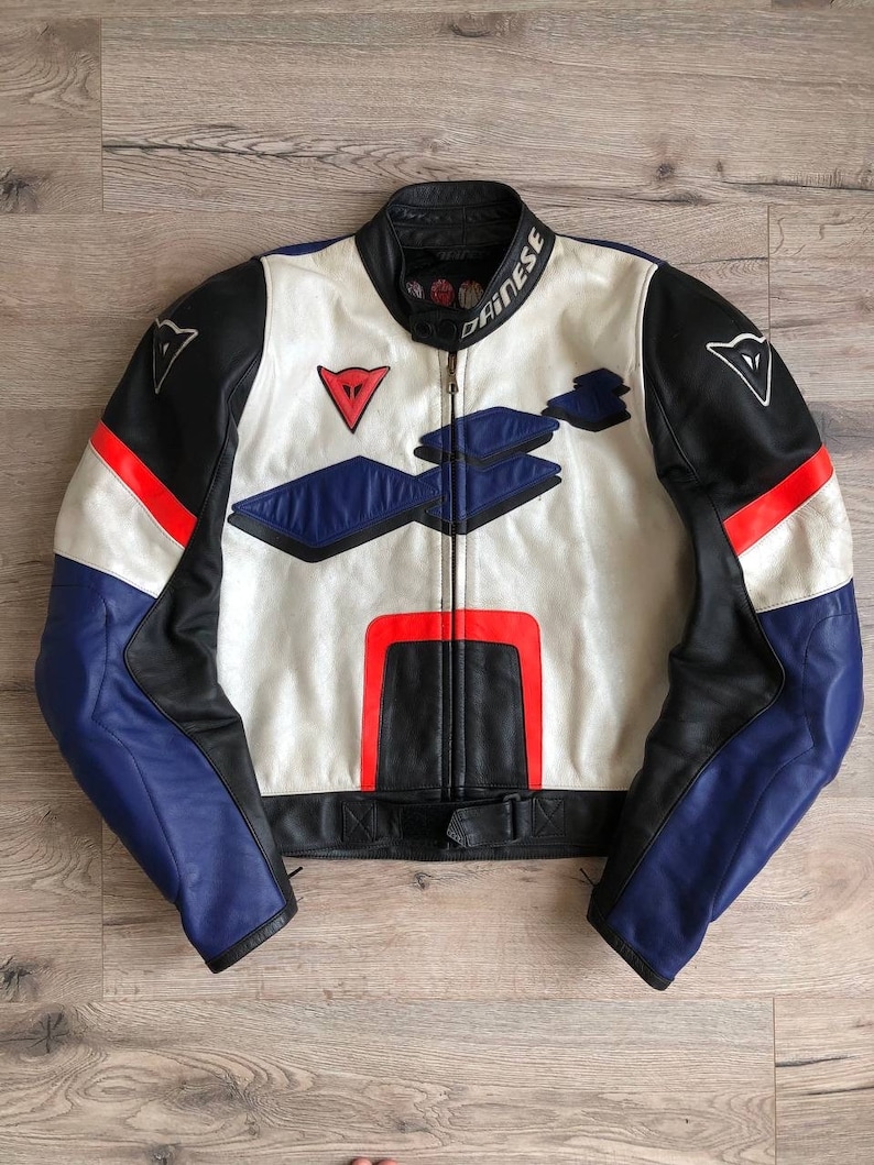 K&ouml;nnte beinhalten: Eine wei&szlig;e, schwarze und blaue Leder-Motorradjacke mit roten Akzenten. Die Jacke hat einen Rei&szlig;verschluss und ein Dainese-Logo auf der linken Schulter und dem rechten &Auml;rmel.