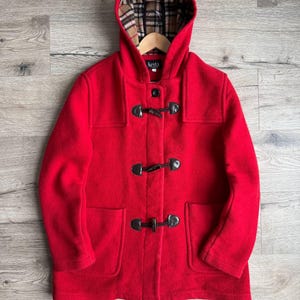 May include: A bright red hooded duffel coat with toggle closures and two front pockets. The hood lining features a classic plaid pattern. The coat is displayed on a wooden hanger against a light wood grain background. The coat has the brand name "kesta" on the label.