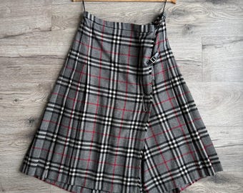 May include: A grey plaid skirt with red and black detailing hangs on a wooden hanger. The skirt has a wrap design with a buckle fastening. The fabric displays a classic tartan pattern.