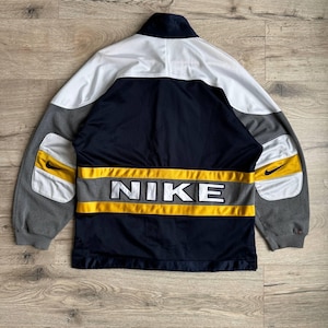 May include: A Nike track jacket featuring a color-blocked design in navy blue, white, grey, and yellow. The back displays the word NIKE in bold white letters across a horizontal yellow and grey stripe, with small black swoosh logos on the sleeves.