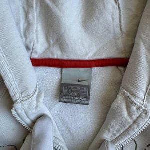 Op de afbeelding: Een wit hooded sweatshirt met een zwarte rits en een rood-wit Nike-label. Het label zegt "US 10, EU 128/140, CN 160, Made in Pakistan, Fabriqu&eacute; au Pakistan."