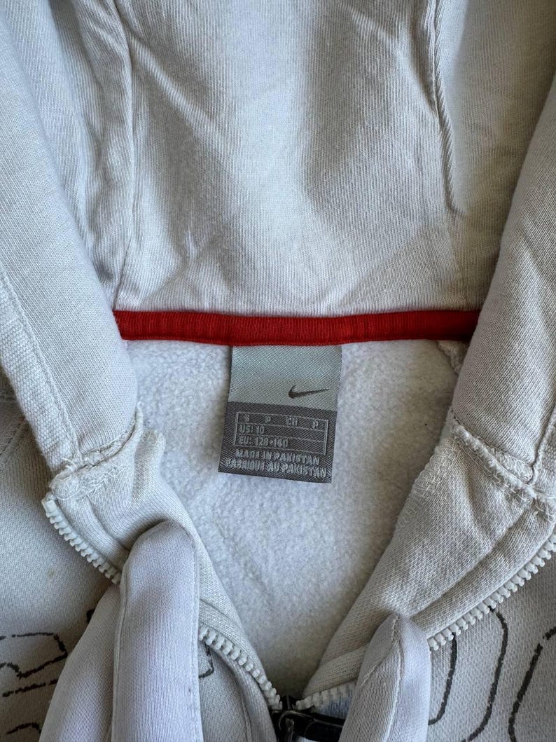 Op de afbeelding: Een wit hooded sweatshirt met een zwarte rits en een rood-wit Nike-label. Het label zegt "US 10, EU 128/140, CN 160, Made in Pakistan, Fabriqu&eacute; au Pakistan."