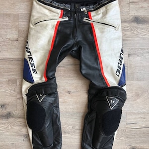K&ouml;nnte beinhalten: Wei&szlig;e und schwarze Leder-Motorradhose mit roten und blauen Akzenten. Die Hose hat Knieschoner und der Markenname "Dainese" ist seitlich aufgedruckt.
