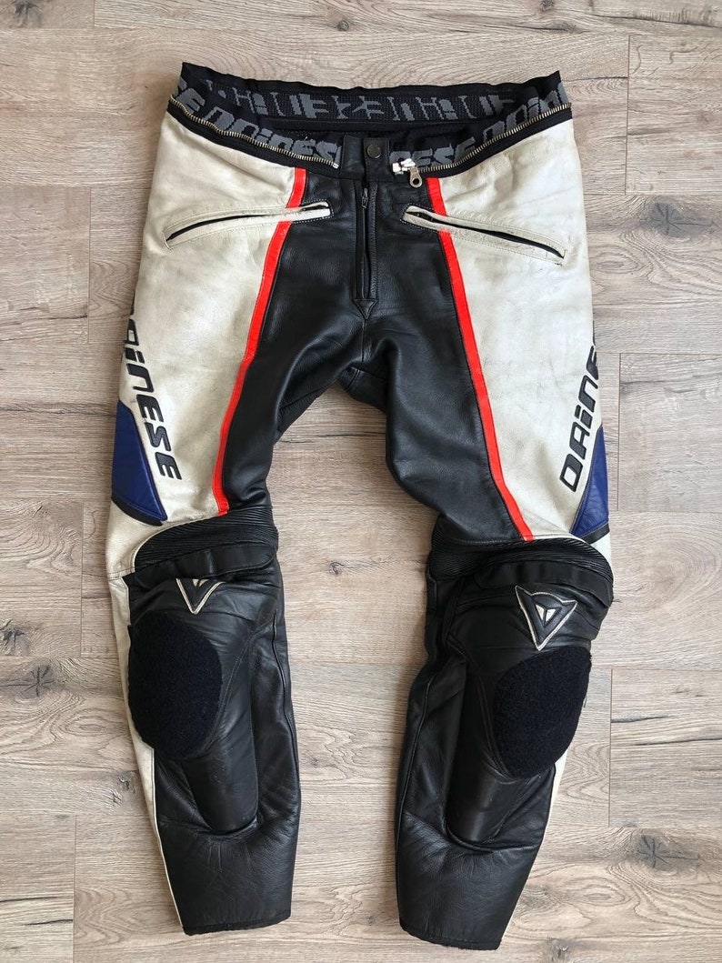 K&ouml;nnte beinhalten: Wei&szlig;e und schwarze Leder-Motorradhose mit roten und blauen Akzenten. Die Hose hat Knieschoner und der Markenname "Dainese" ist seitlich aufgedruckt.