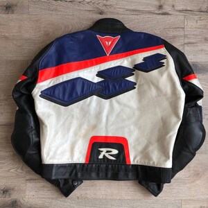 K&ouml;nnte beinhalten: Eine wei&szlig;e, schwarze, blaue und rote Leder-Motorradjacke mit einem Dainese-Logo auf der R&uuml;ckseite. Die Jacke hat ein rotes "R" auf der R&uuml;ckseite.