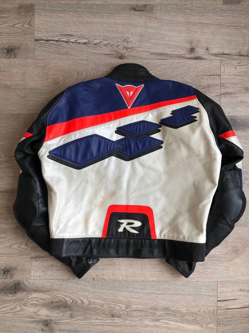 K&ouml;nnte beinhalten: Eine wei&szlig;e, schwarze, blaue und rote Leder-Motorradjacke mit einem Dainese-Logo auf der R&uuml;ckseite. Die Jacke hat ein rotes "R" auf der R&uuml;ckseite.