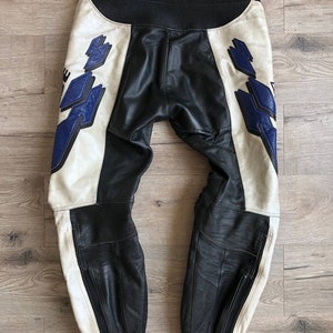 K&ouml;nnte beinhalten: Schwarze und wei&szlig;e Leder-Motorradhose mit blauen Akzenten und einem Rei&szlig;verschlussdetail. Die Hose hat einen wei&szlig;en Streifen an der Seite jedes Beins.