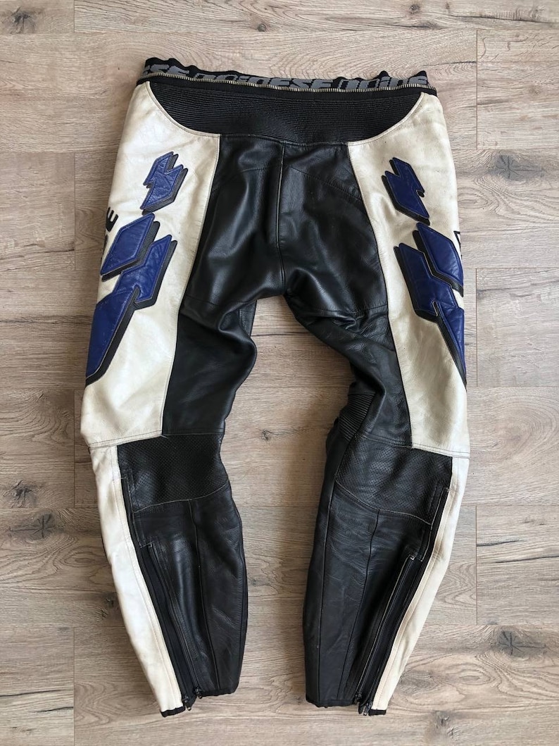 K&ouml;nnte beinhalten: Schwarze und wei&szlig;e Leder-Motorradhose mit blauen Akzenten und einem Rei&szlig;verschlussdetail. Die Hose hat einen wei&szlig;en Streifen an der Seite jedes Beins.