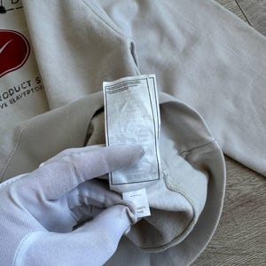 Op de afbeelding: Een lichtgrijs sweatshirt met een rood swoosh-logo en de tekst "Product of Beaverton, OR" op de voorkant. Het sweatshirt heeft een label met de tekst "0651Y 1425" erop.