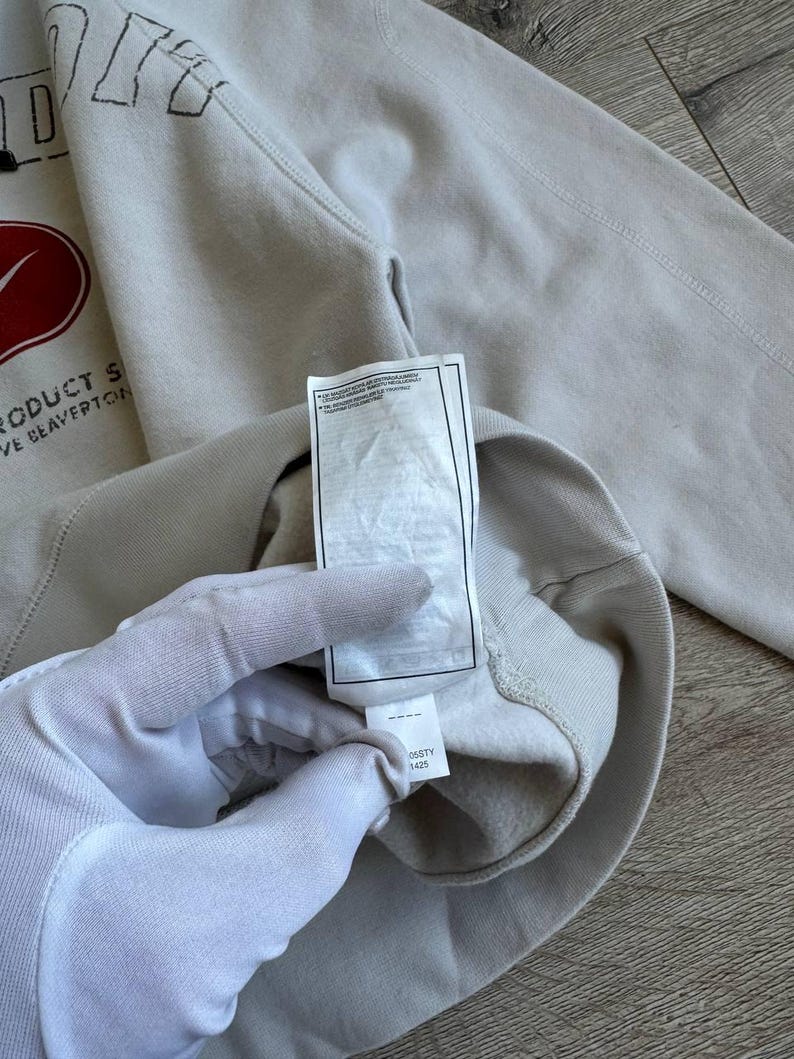 Op de afbeelding: Een lichtgrijs sweatshirt met een rood swoosh-logo en de tekst "Product of Beaverton, OR" op de voorkant. Het sweatshirt heeft een label met de tekst "0651Y 1425" erop.