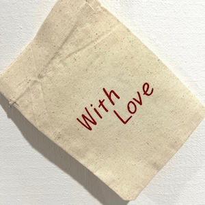 May include: A small, beige canvas bag with the words "With Love" printed in red. The bag is made of a natural-colored fabric and is slightly angled, showing its texture and the simple, heartfelt message.