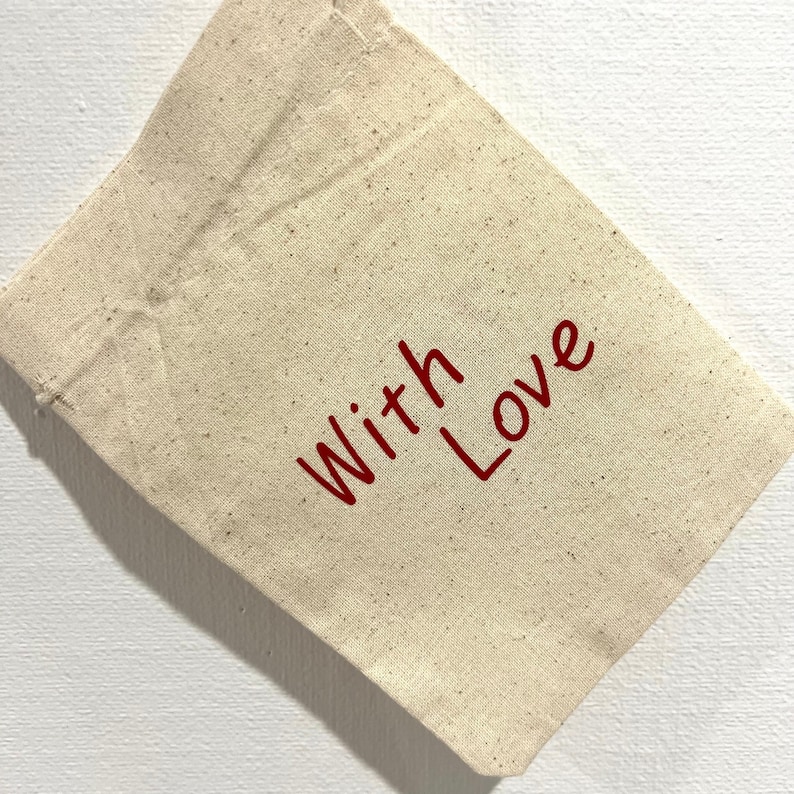 May include: A small, beige canvas bag with the words "With Love" printed in red. The bag is made of a natural-colored fabric and is slightly angled, showing its texture and the simple, heartfelt message.