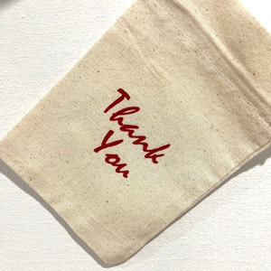 May include: A small, beige canvas bag with the words "Thank You" printed in red script. The bag is folded over, and the stitching is visible along the top edge. The background is a white surface.