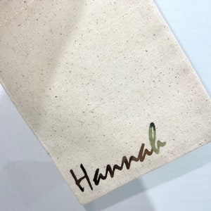May include: A beige canvas bag with the name "Hannah" printed in a metallic, cursive font. The bag has a simple, minimalist design, with a slightly textured surface.