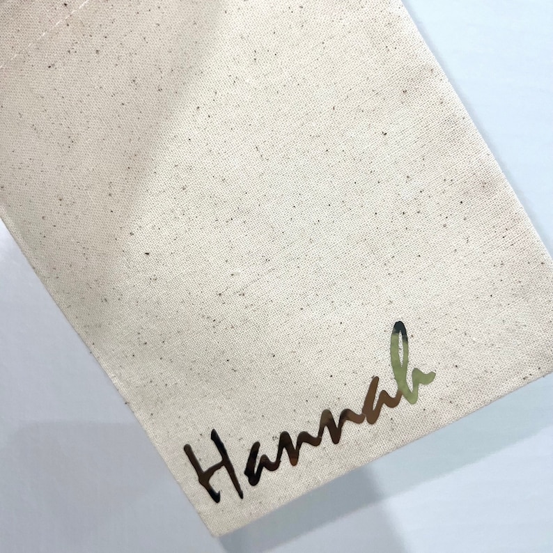 May include: A beige canvas bag with the name "Hannah" printed in a metallic, cursive font. The bag has a simple, minimalist design, with a slightly textured surface.