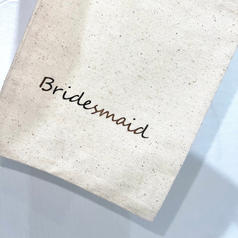 May include: A beige canvas tote bag with the word "Bridesmaid" printed in a dark brown script. The bag has a simple, minimalist design, perfect for wedding favors or gifts.