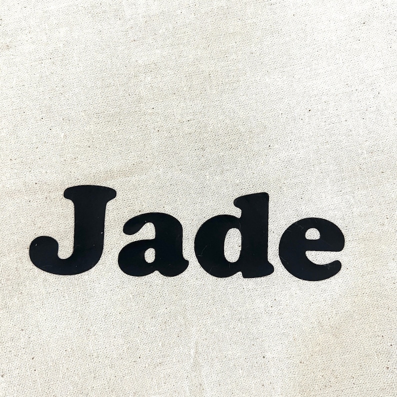 May include: Close-up of a beige fabric with the name "Jade" printed in bold, black letters. The letters have a rounded, playful font. The fabric has a slightly textured appearance.