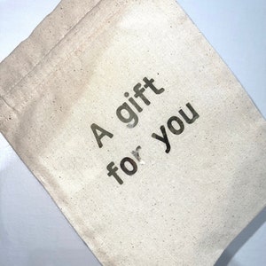 May include: A beige canvas gift bag with the words "A gift for you" printed in dark gray. The bag is made of a natural, textured fabric and has a simple, drawstring closure.