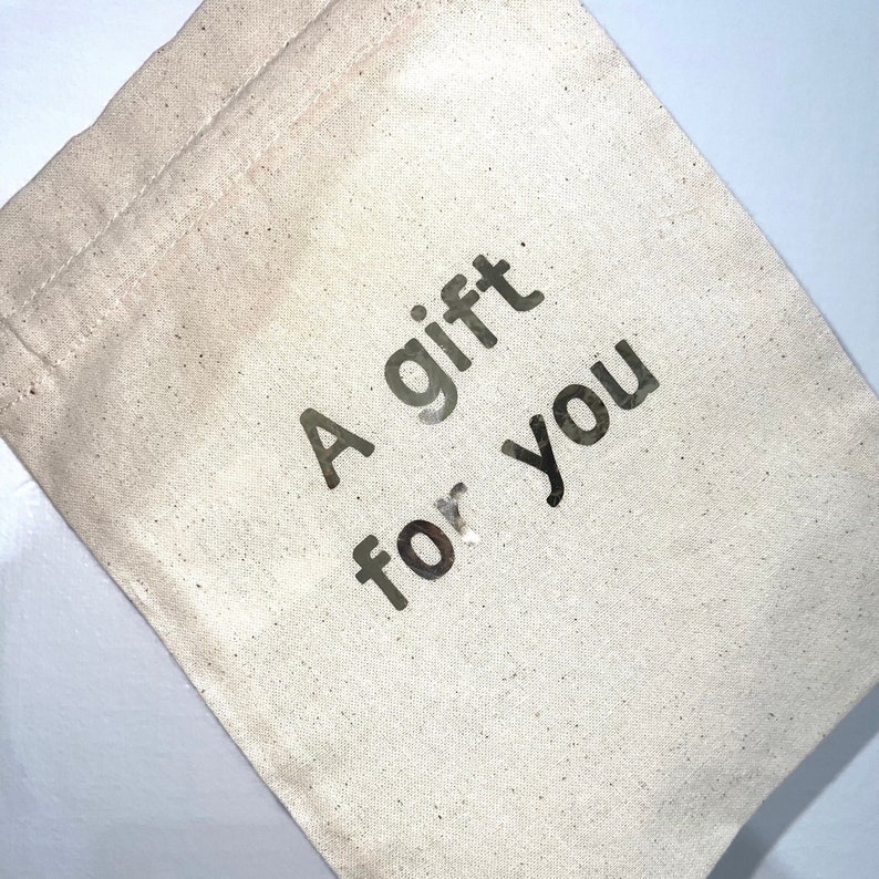 May include: A beige canvas gift bag with the words "A gift for you" printed in dark gray. The bag is made of a natural, textured fabric and has a simple, drawstring closure.