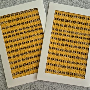 May include: Two framed art prints. Each print features a yellow and black woven pattern within a white border. The textured pattern is a repeating series of small, dark dots and lines against a bright yellow background. The frames are rectangular.