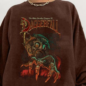 May include: A brown sweatshirt featuring a graphic from "The Elder Scrolls Chapter II: Daggerfall." The design includes a skull with a sword and clawed hands. The text "Daggerfall" is in red and gold.