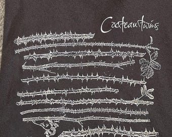 May include: Dark gray t-shirt with a white graphic of thorny vines and the text "Cocteau Twins". The shirt is folded and appears to be made of a soft material. The design is centered on the front of the shirt.