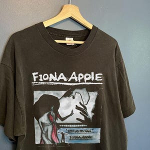 Może przedstawiać: Czarny T-shirt z białym nadrukiem okładki albumu Fiony Apple "Fast as You Can". Okładka albumu przedstawia czarno-białą ilustrację kobiety w czerwonej sukience.