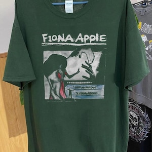 Może przedstawiać: Zielony T-shirt z białym nadrukiem graficznym przedstawiającym okładkę albumu Fiony Apple "When the Pawn...". Okładka albumu przedstawia czarno-białą ilustrację kobiety w czerwonej sukience, do kt&oacute;rej wyciągnięta jest ręka.