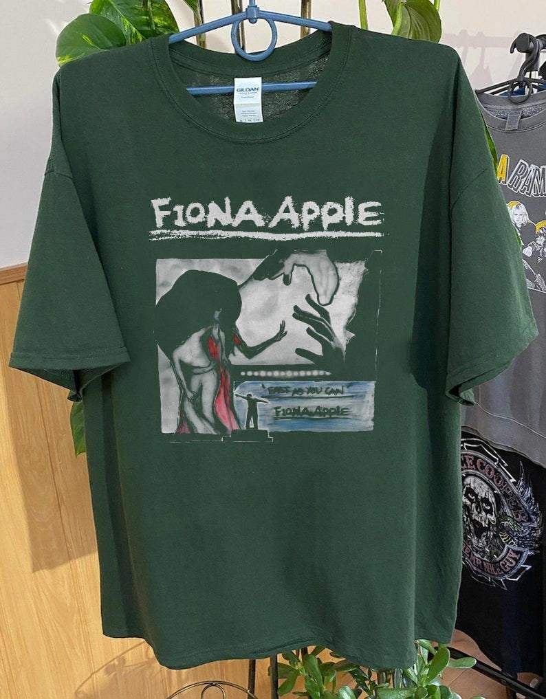 Może przedstawiać: Zielony T-shirt z białym nadrukiem graficznym przedstawiającym okładkę albumu Fiony Apple "When the Pawn...". Okładka albumu przedstawia czarno-białą ilustrację kobiety w czerwonej sukience, do kt&oacute;rej wyciągnięta jest ręka.