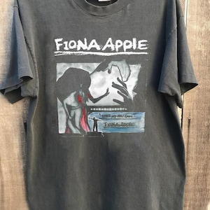 Może przedstawiać: Czarny T-shirt z nadrukiem graficznym Fiona Apple. Grafika przedstawia czarno-biały obraz kobiety w czerwonej sukience i mężczyzny stojącego na scenie. Tekst "This is as you can" i "Fiona Apple" jest nadrukowany na grafice.