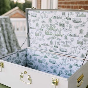 May include: A white rectangular storage trunk with gold-toned metal latches and handles. The interior is lined with white fabric featuring a repeating green illustrative pattern of city landmarks, trees, and monograms, including the text The Bransides 2025.