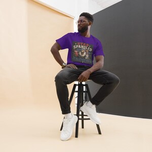 May include: A person wearing a purple t-shirt with the words "Star Spangled" above an eagle graphic. The graphic includes red, white, and blue elements. The person is wearing black jeans and white sneakers, sitting on a black stool.