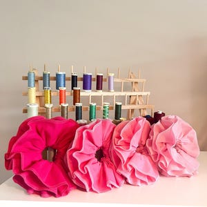 May include: Four large, ruffled scrunchies in shades of pink sit on a white surface. Behind them, a wooden thread rack displays spools of colourful thread. The scrunchies have a circular design with a ruffled edge.