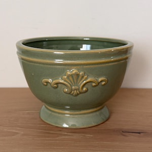 Può includere: Una ciotola in ceramica verde con base a piedistallo e decorativo motivo floreale in rilievo. La ciotola ha un'ampia apertura e un bordo leggermente svasato. L'esterno è verde opaco con un accento dorato attorno al bordo e al motivo in rilievo.