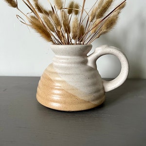 Op de afbeelding: Een handgemaakte keramische vaas met een handvat, met een gespikkelde off-white en bruine kleurstelling. De vaas is gevuld met gedroogde, beige siergrassen. De vaas heeft een unieke, afgeronde vorm en een stevig handvat.