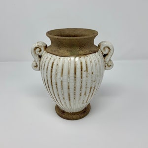 Może przedstawiać: Biały ceramiczny wazon z brązową obręczą i podstawą, z pionowymi żebrowanymi detalami. Wazon ma dwa ozdobne uchwyty i postarzaną, spękaną powierzchnię. Ten dekoracyjny wazon nadaje się do dekoracji wnętrz. (wysokość ok. 20 cm)